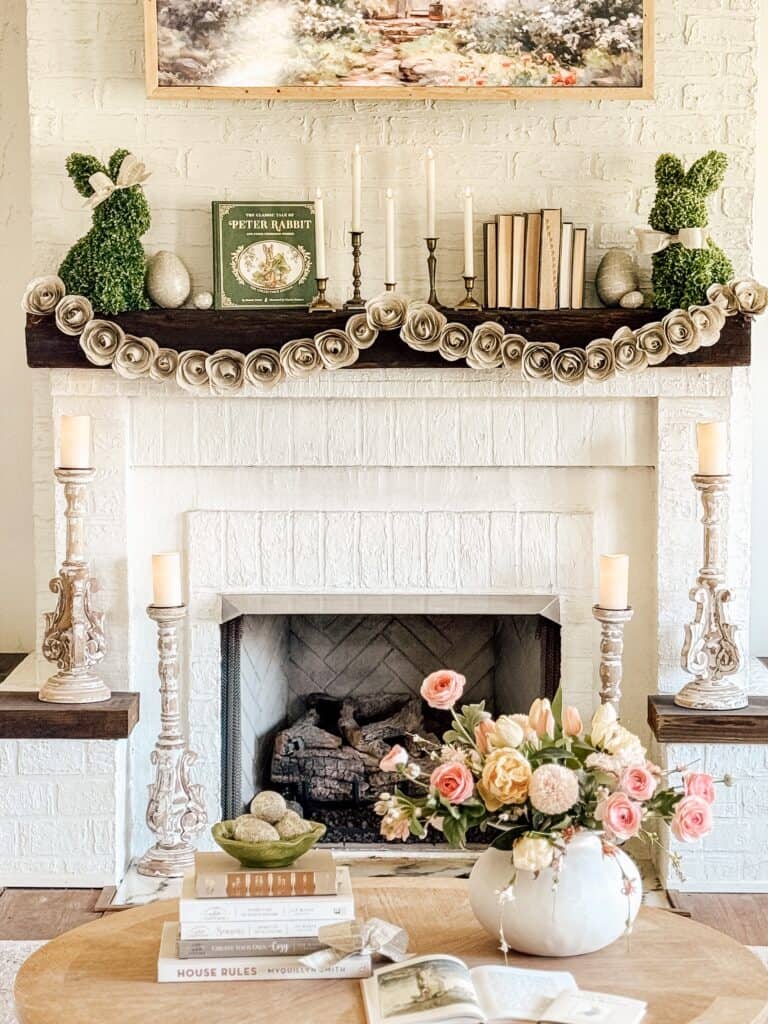 A cozy living room with a white brick fireplace decorated with greenery, candles, and a Peter Rabbit book. A round wooden table holds a vase of pink and cream flowers. Stacked books and tall candle holders complete the warm and inviting scene.