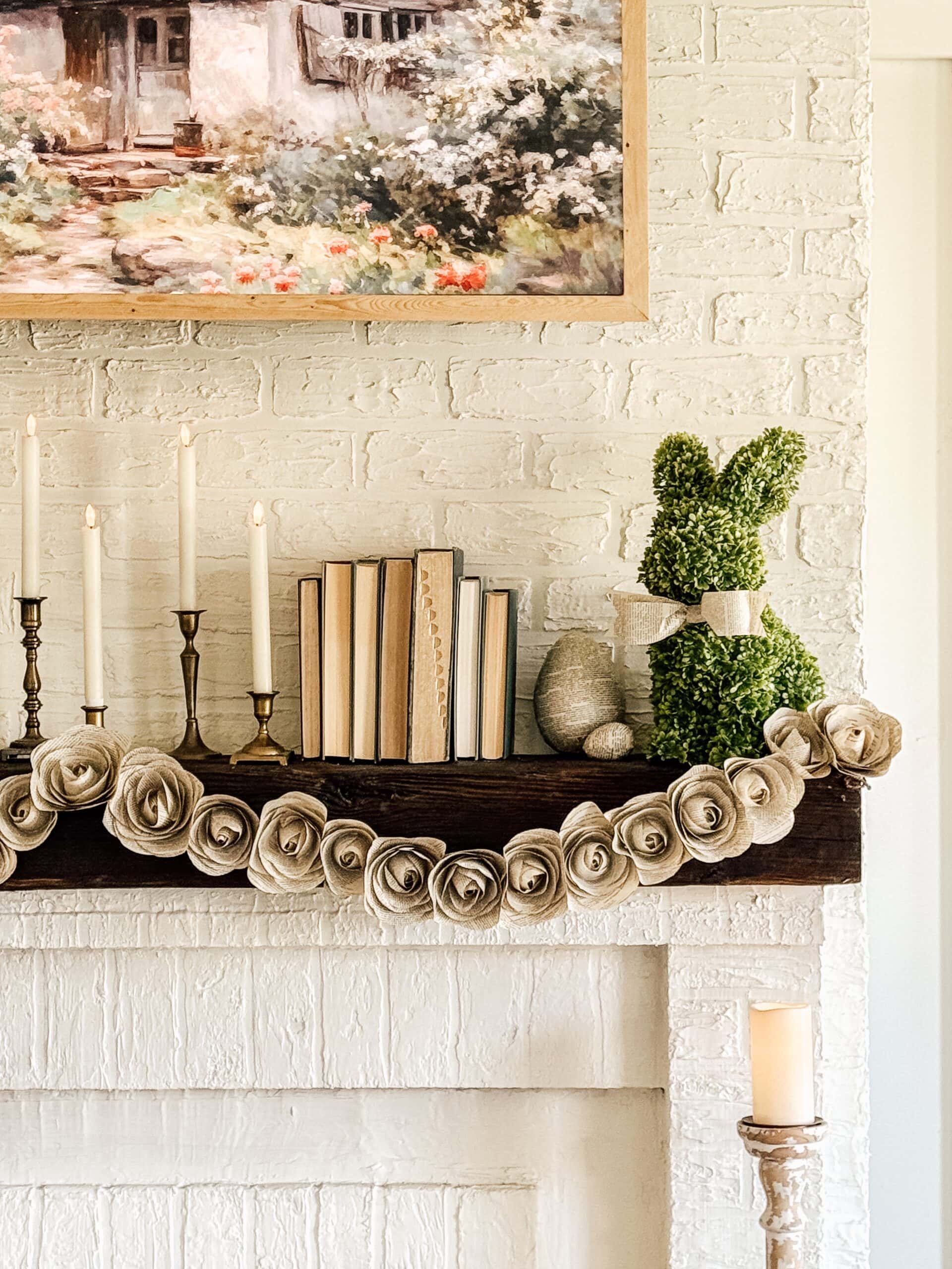 A rustic mantel decorated with books, candles, and a greenery bunny sculpture. A garland of fabric roses drapes across the mantel. A painting of a cottage scene hangs above, all set against a white brick wall. A lit candle stands nearby.