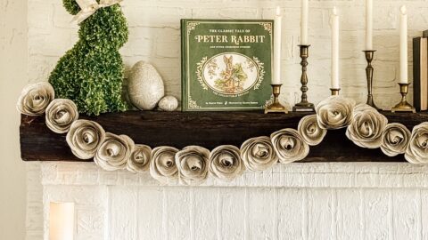 A white brick fireplace decorated with a green moss rabbit figure, a Peter Rabbit book, a stone egg, and candlesticks on the mantel. A garland of textured flowers drapes across the mantel. Lit candles stand on ornate holders nearby.