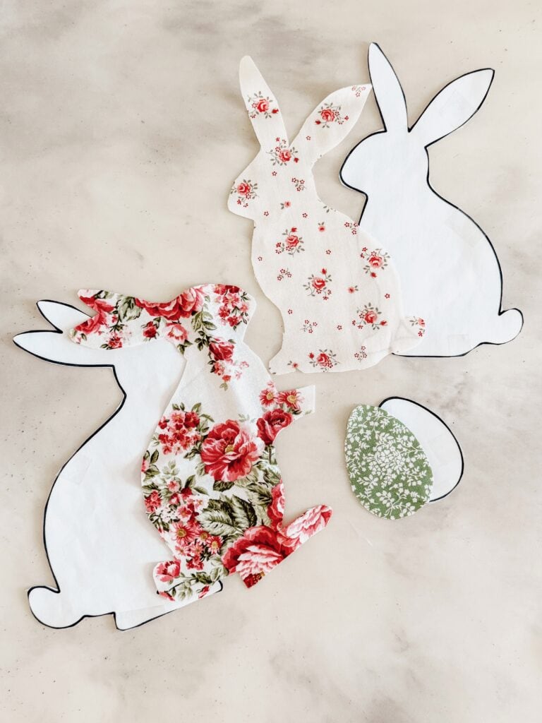 Cut-out shapes of rabbits and an egg, made from floral-patterned and plain white fabric, are arranged on a light, textured surface. The designs give a spring or Easter-themed craft vibe.
