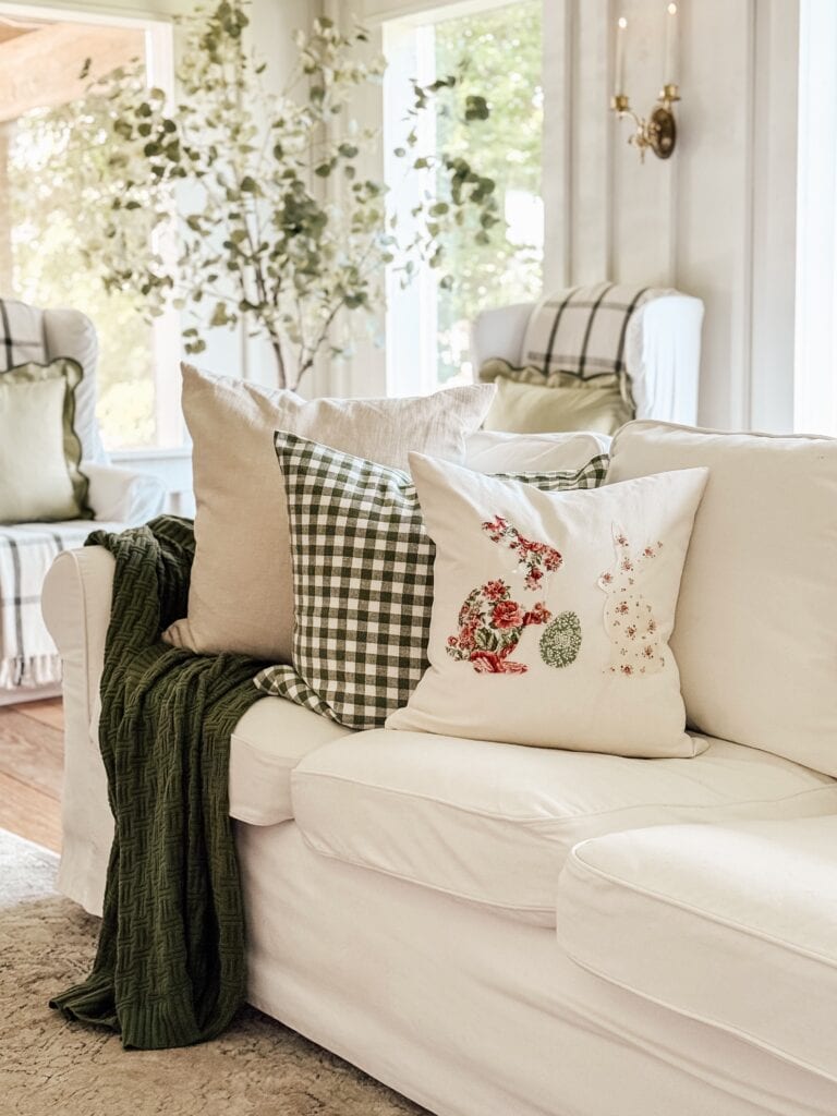 A white sofa with pillows in green gingham, beige, and embroidered floral patterns, draped with a dark green blanket, sits in a bright, airy living room with plants and large windows in the background.