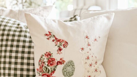 A decorative pillow on a white couch features embroidered silhouettes of two rabbits and an egg, adorned with floral patterns. Behind it, a green and white checkered pillow complements the cozy, fresh atmosphere of the room.