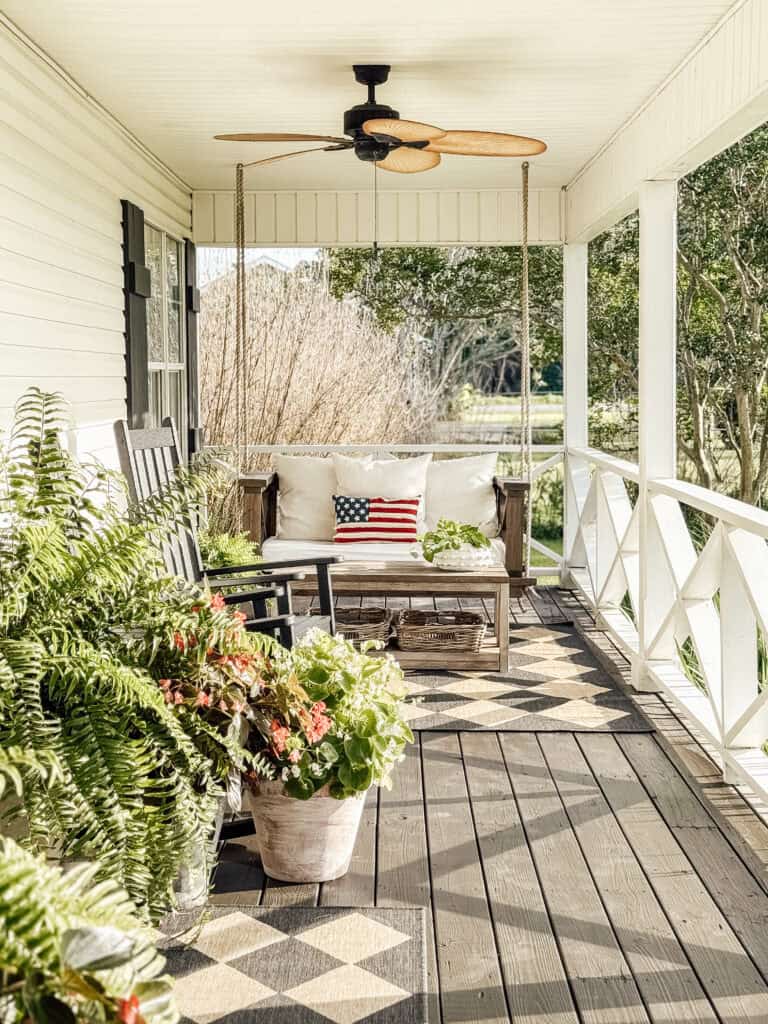 A cozy porch with potted plants, black rocking chairs, a wooden swing with beige cushions, and an American flag pillow. A ceiling fan hangs above, and the porch overlooks a green, sunny yard.