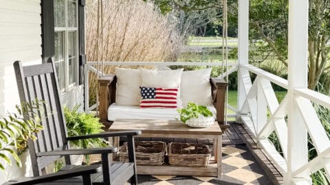 A cozy front porch with a wooden swing, an American flag pillow, a black rocking chair, green potted plants, a ceiling fan, and a geometric-patterned rug on a wooden floor.