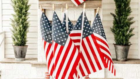 A patriotic centerpiece with small American flags and glass soda bottles sits on a wooden table. In the background, a grapevine wreath with an American flag hangs above the fireplace, framed by two potted evergreen trees.