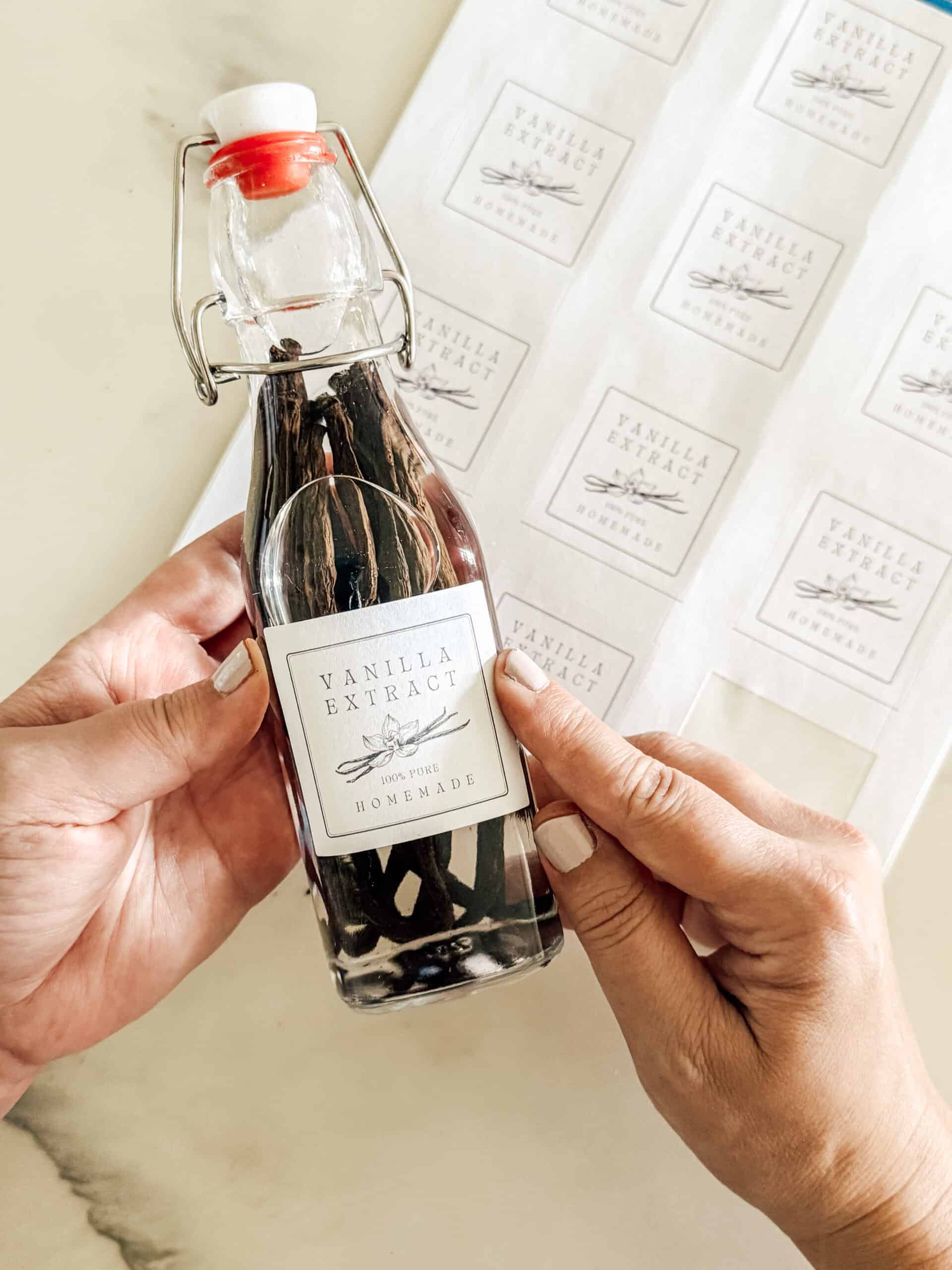 A person holds a glass bottle of homemade vanilla extract with a white label, next to a sheet of similar labels on a marble surface. The bottle contains dark vanilla beans and has a swing-top cap.