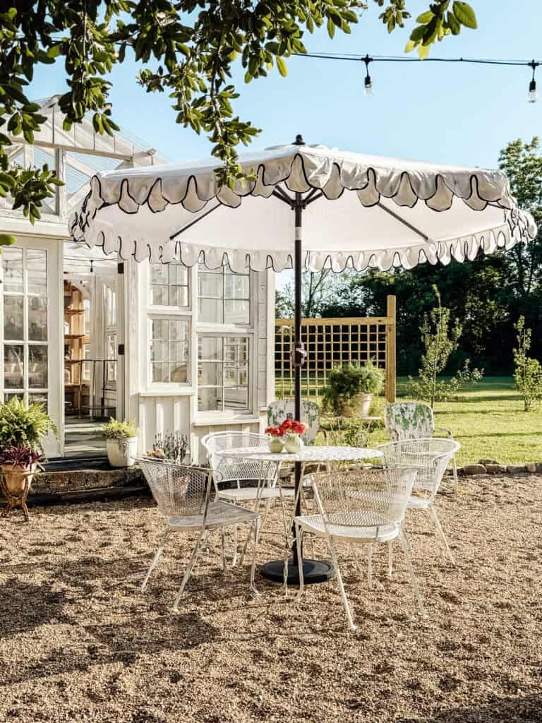 A white metal outdoor table with four matching chairs sits under a ruffled white umbrella on a gravel patio next to a glass greenhouse, with plants and greenery in the background.