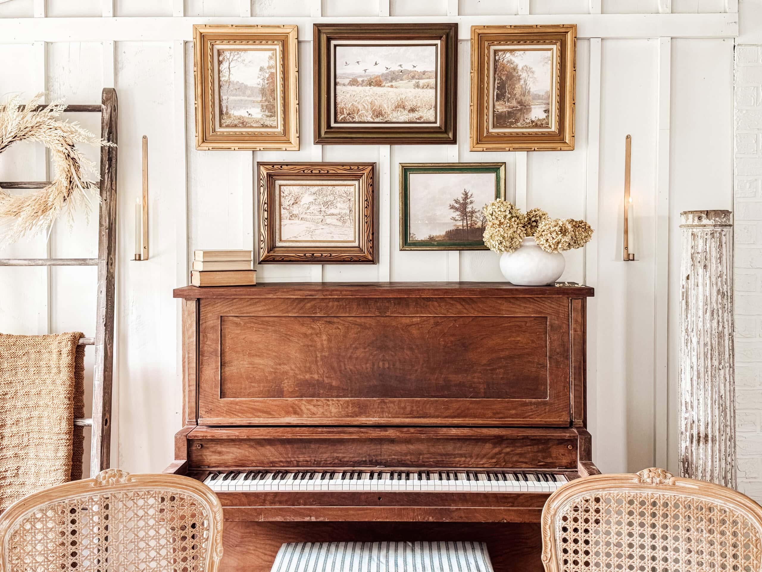 A vintage upright piano sits against a white paneled wall, topped with books and a vase of dried flowers. Above, digital vintage art and landscape prints are arranged gallery style. Cane-backed chairs are in the foreground.
