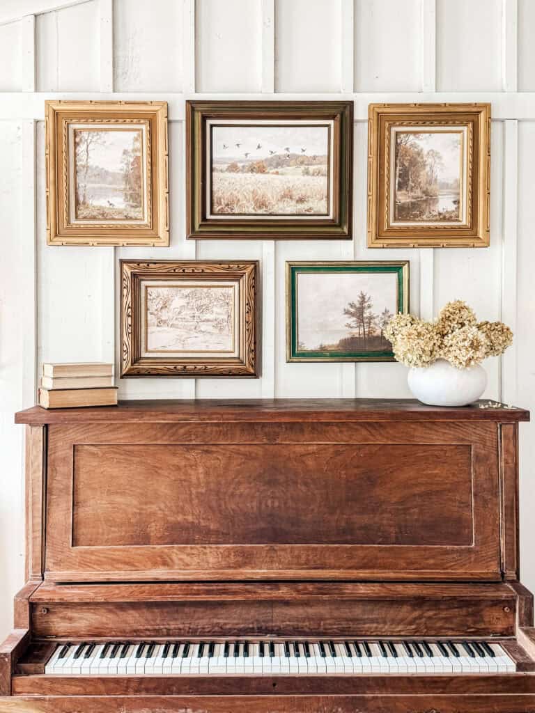 A wooden upright piano with white and black keys stands against a white paneled wall. On top are stacked books and a vase of dried flowers. Above the piano, five framed digital vintage art landscape paintings hang in a neat row.