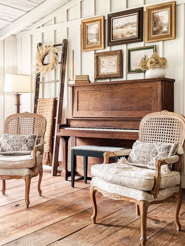 A cozy vintage living room with two ornate armchairs, a wooden piano, framed digital vintage art on white paneled walls, a ladder with a wreath, and natural wood floors, creating a warm, rustic atmosphere.