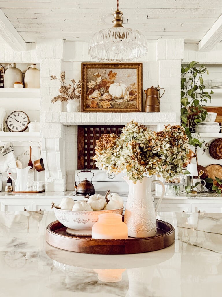 A bright, cozy kitchen with white decor features a tray on a marble counter holding a ceramic pitcher of dried flowers, a bowl of white pumpkins, and a lit candle; shelves and autumn-themed art are in the background.