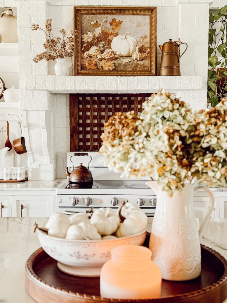 A cozy, vintage-style kitchen with white decor, dried flowers in a vase, small white pumpkins in a bowl, a lit candle, and a painting of pumpkins and leaves above the stove on a white brick wall.