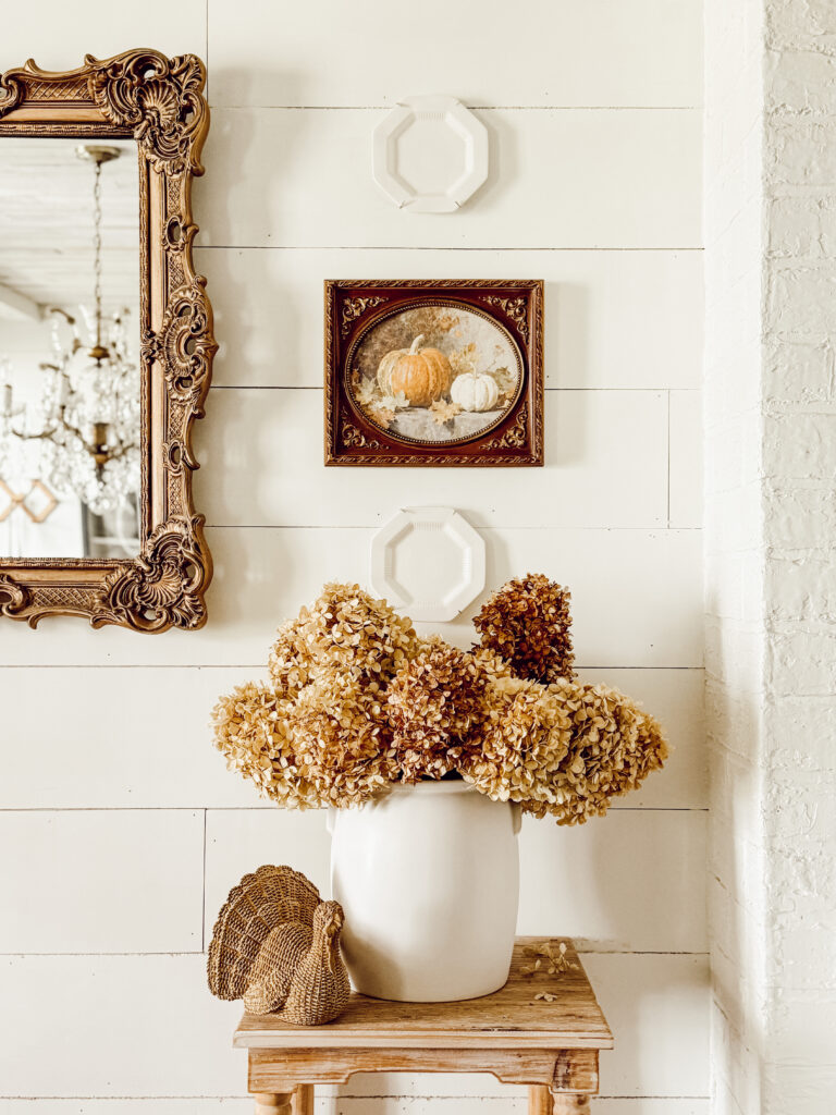 A white vase with dried hydrangeas sits on a wooden table beside a decorative woven turkey. Above, a framed painting of pumpkins and two empty white frames hang on a white shiplap wall next to an ornate mirror.