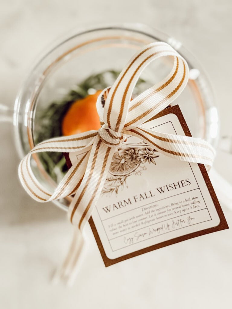 A clear glass mug with a striped cream and gold ribbon tied around it, holding a card that reads Warm Fall Wishes and an orange inside the mug, set on a light marble surface.