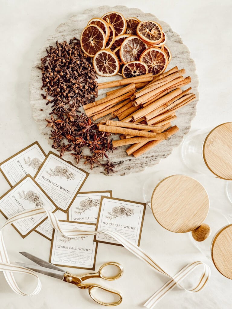 A round tray with dried orange slices, cinnamon sticks, star anise, and cloves. Nearby are jar labels, white ribbon, a gold pair of scissors, and jars with wooden lids on a light surface.