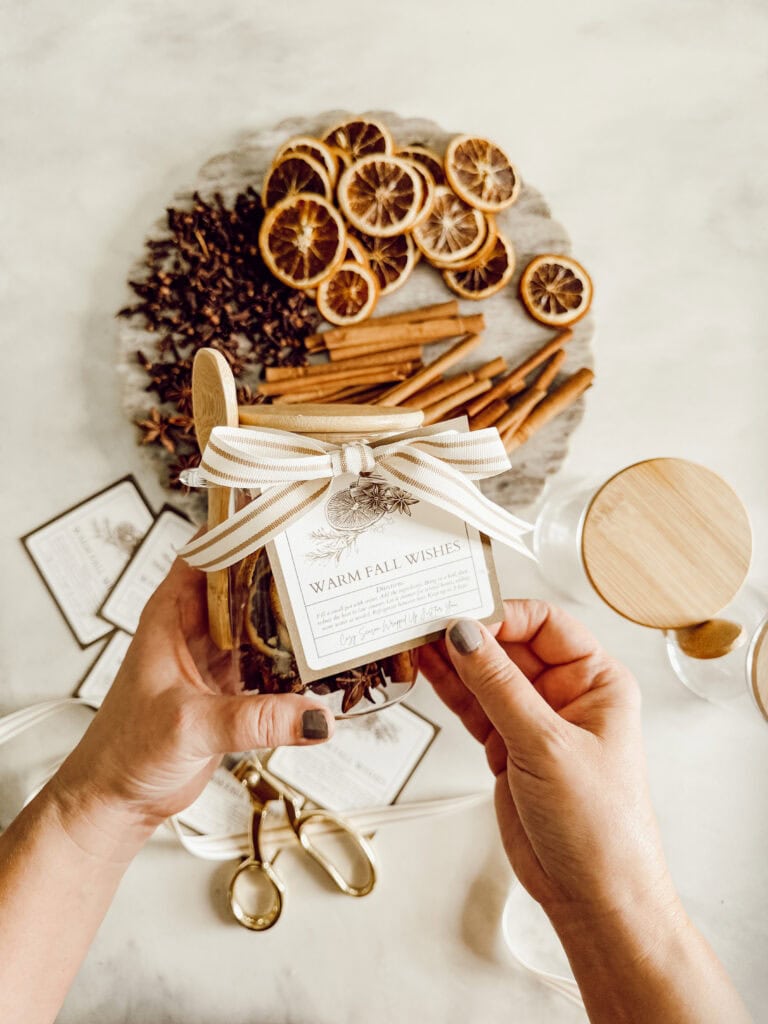 Hands holding a jar with a “Warm Fall Wishes” label and a ribbon. In the background, there’s a tray with dried orange slices, cinnamon sticks, and star anise, along with scissors and ribbon on a light surface.