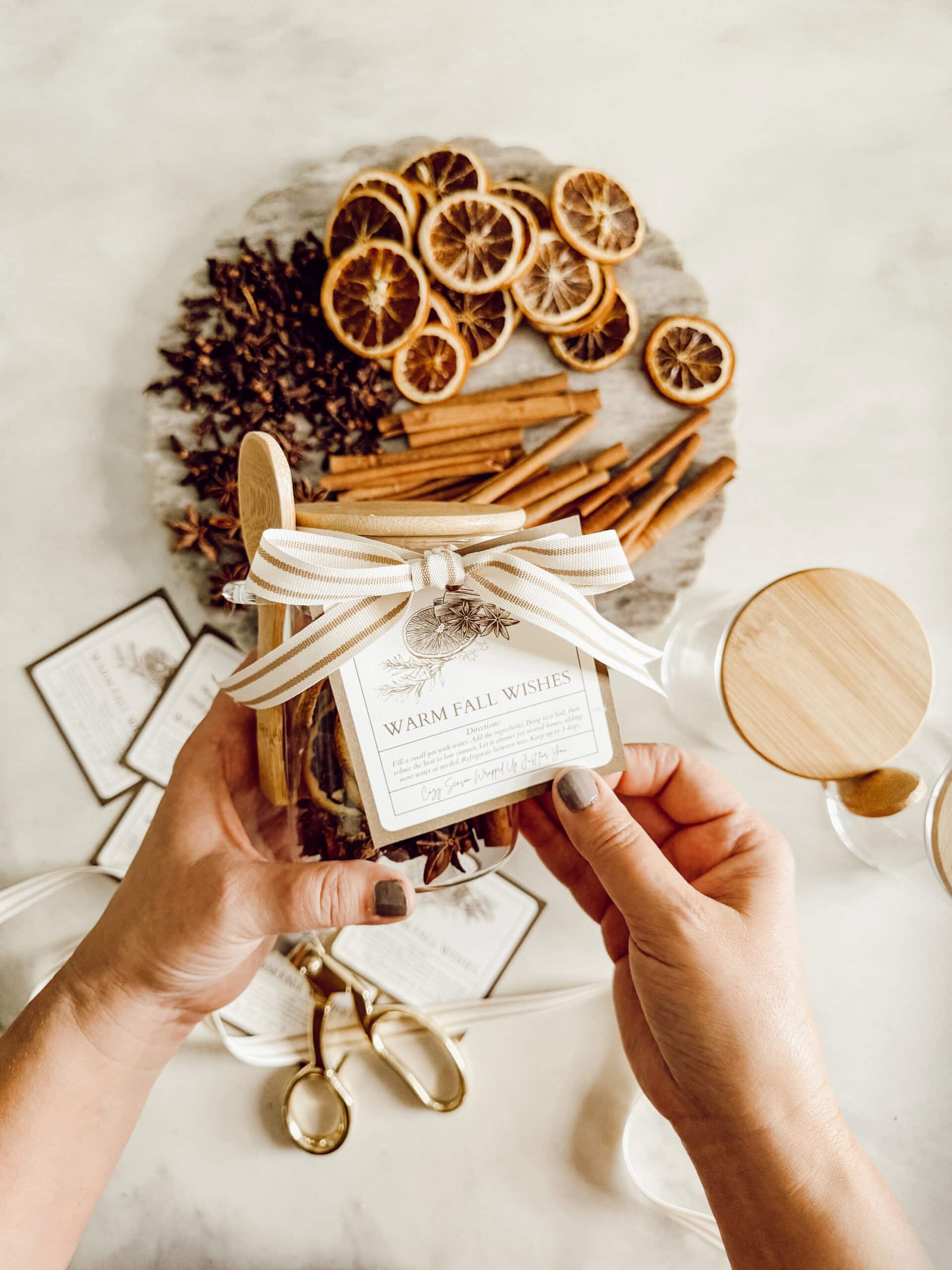 Hands holding a jar with a “Warm Fall Wishes” label and a ribbon. In the background, there’s a tray with dried orange slices, cinnamon sticks, and star anise, along with scissors and ribbon on a light surface.