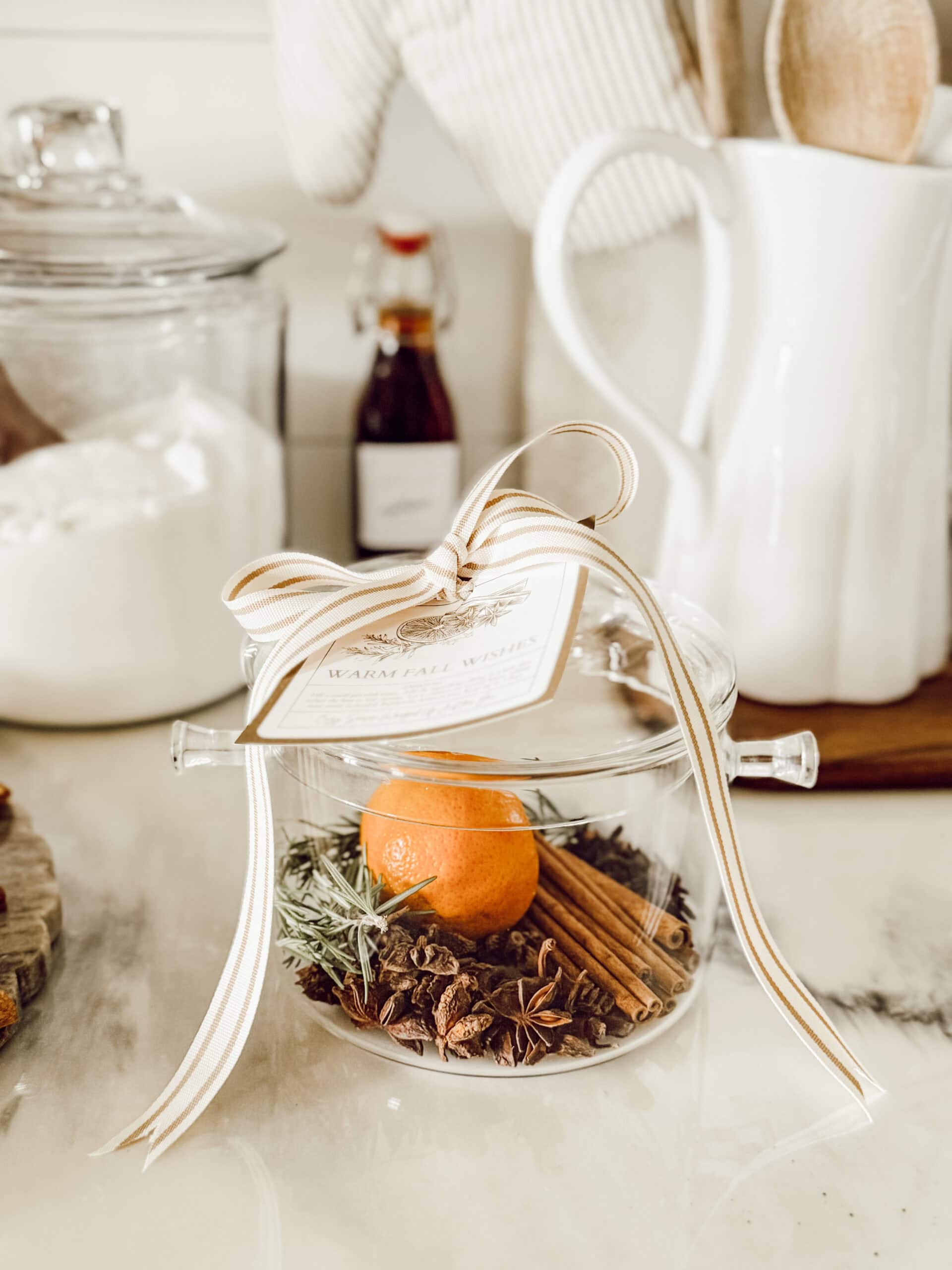 A glass jar filled with a small orange, cinnamon sticks, star anise, and herbs, topped with a striped ribbon and a tag, sits on a kitchen counter beside a sugar jar and kitchen utensils.