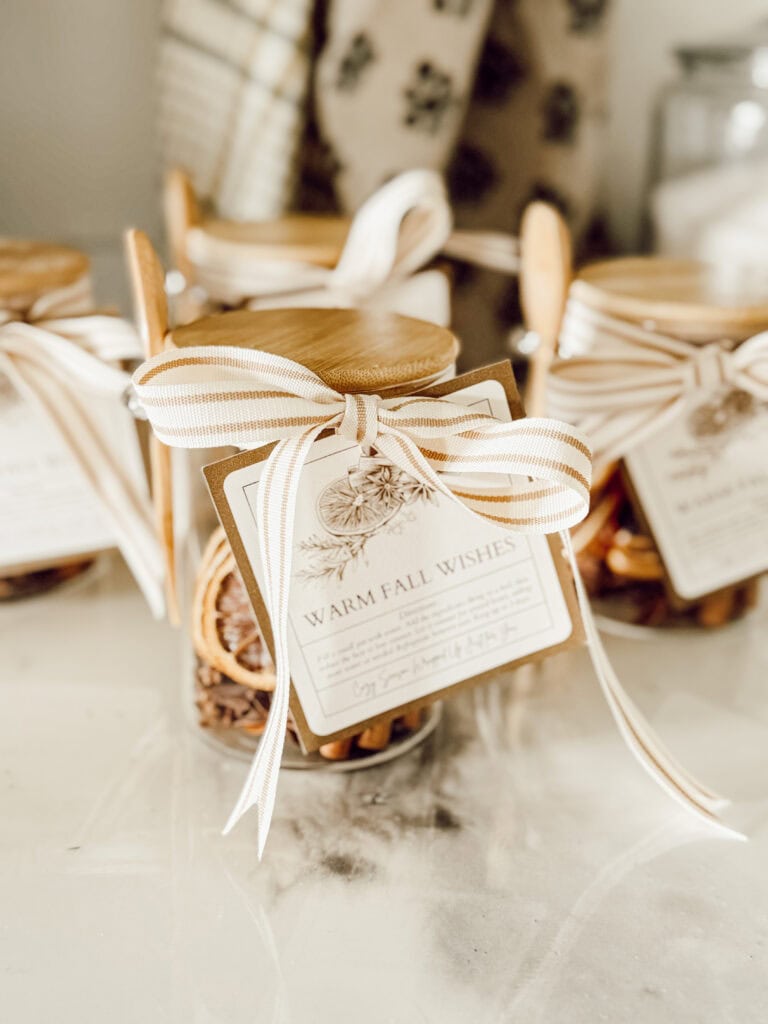 A close-up of glass jars filled with dried orange slices and spices, each tied with a cream ribbon and a “Warm Fall Wishes” tag, arranged on a light-colored surface.