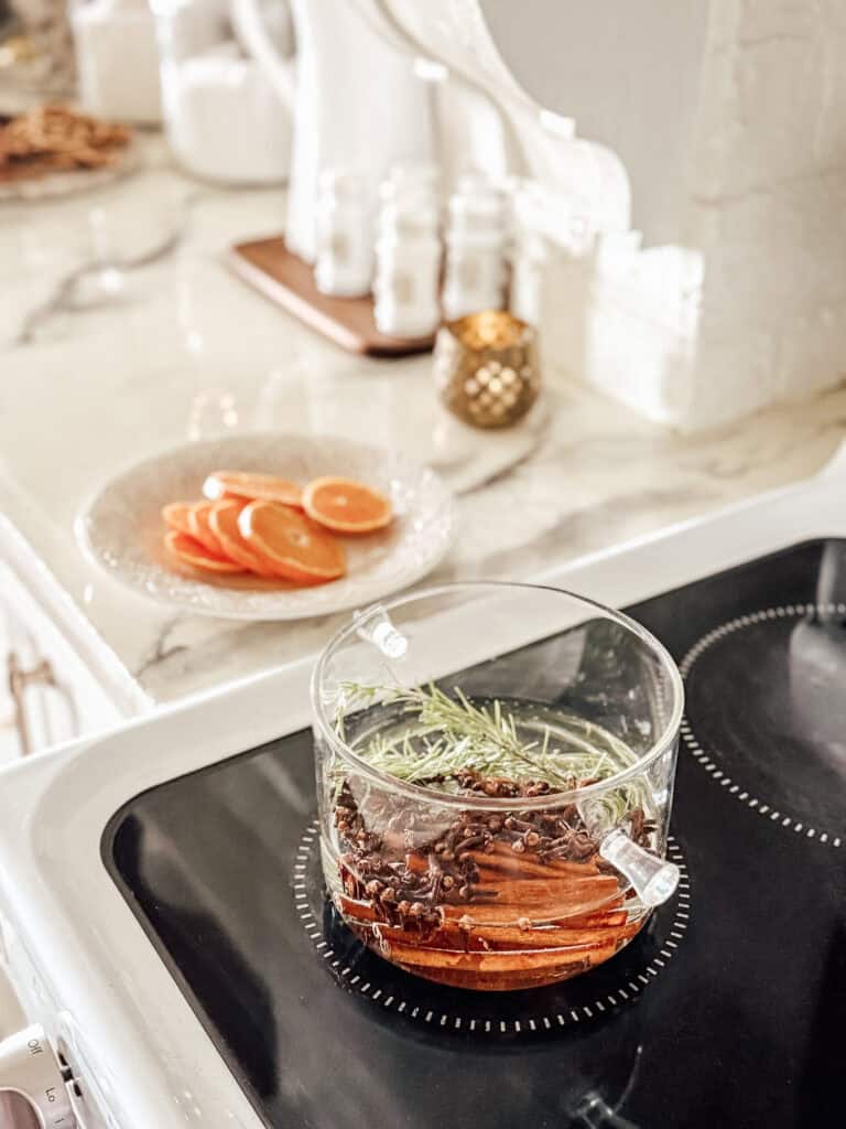 A glass pot with sliced oranges, rosemary, and spices simmers on a stovetop; a plate of orange slices and kitchen decor are visible in the background.