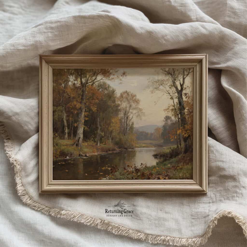 A framed digital vintage art landscape of a tranquil river surrounded by autumn trees rests on beige, textured fabric with fringed edges. The scene captures soft light and muted fall colors.