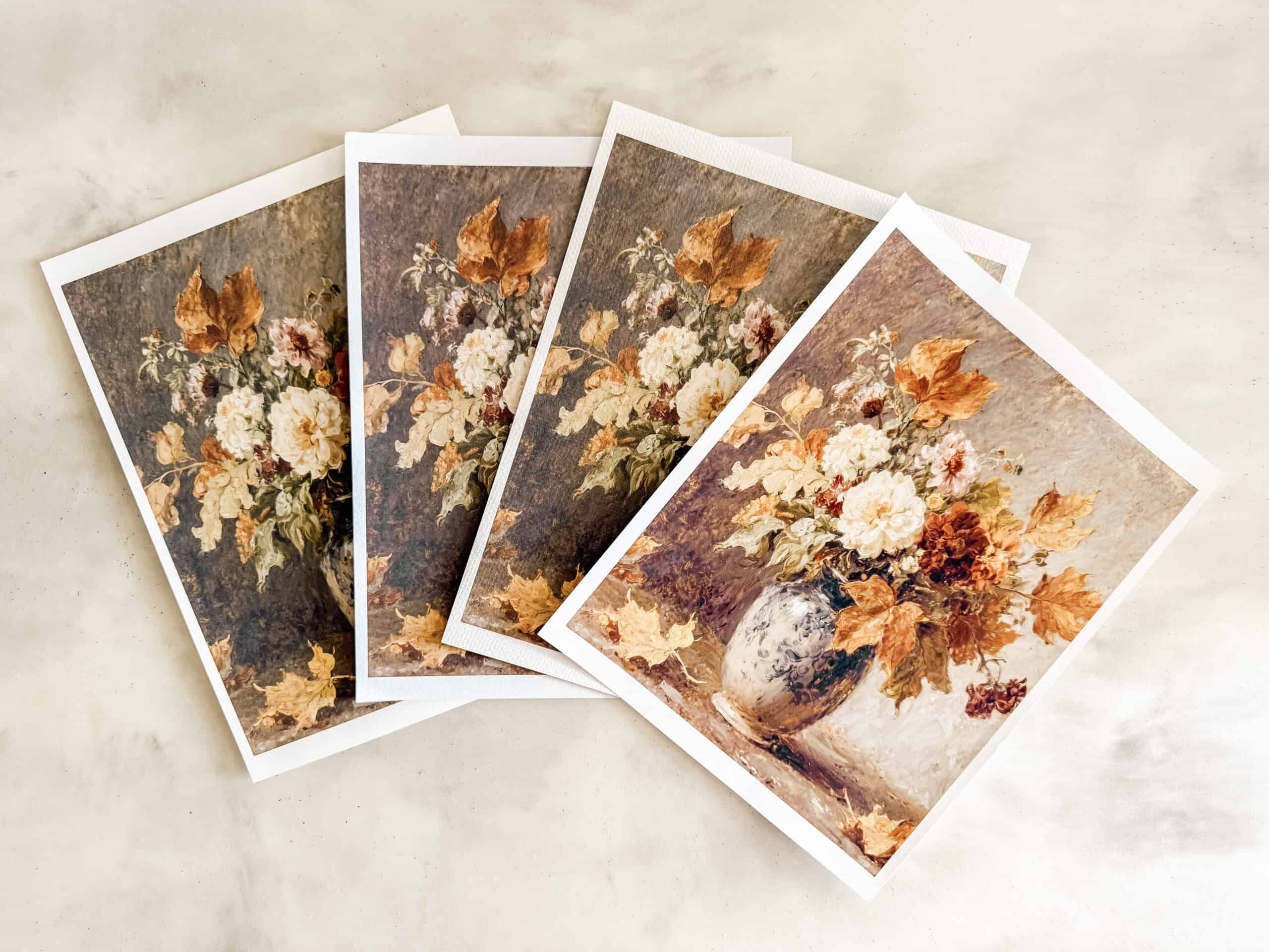 Four greeting cards featuring a painting of a floral bouquet in a vase with autumn-colored leaves, arranged in a fan shape on a light surface.