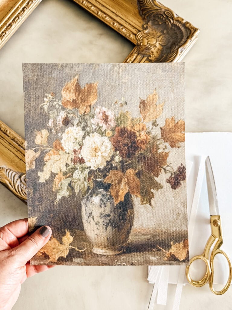 A hand holds a print of a classic still life painting showing a vase with white and brown flowers. Nearby are gold scissors, white papers, and an ornate empty gold frame on a light surface.