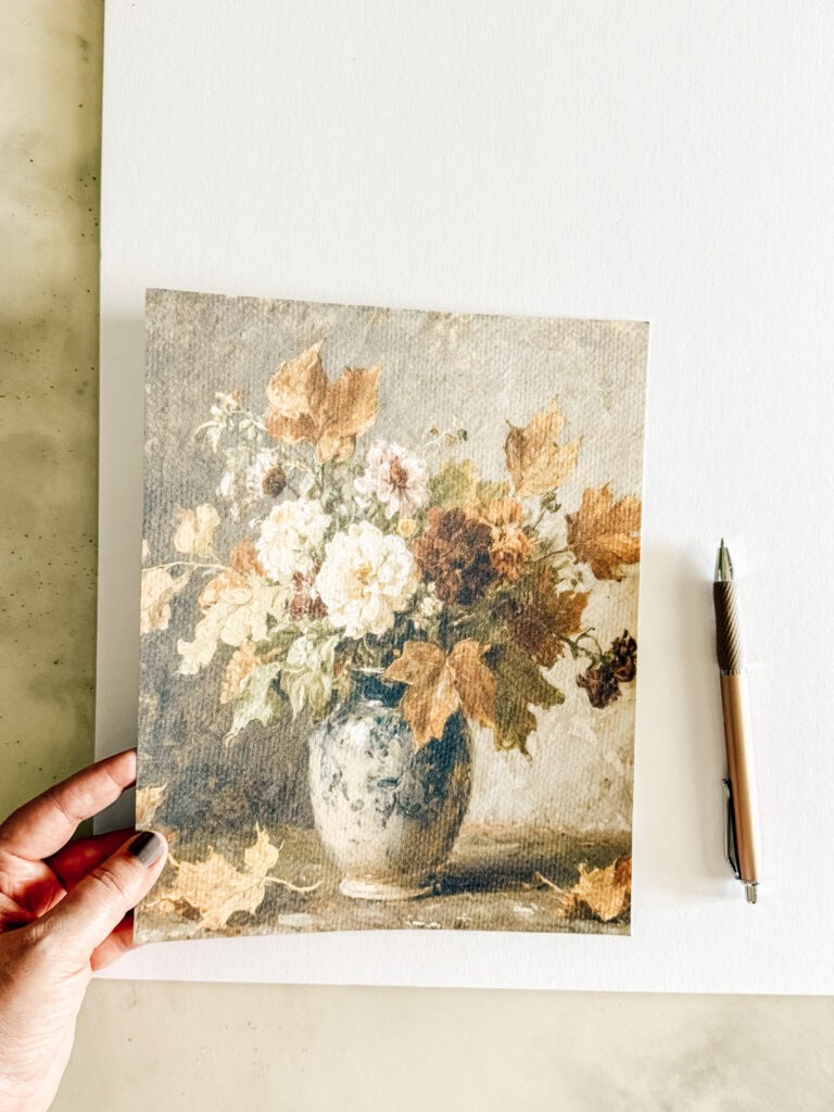A hand holds a painting of a floral arrangement in a blue vase with autumn leaves, placed on a blank white sheet. A pen lies to the right of the artwork on the table.