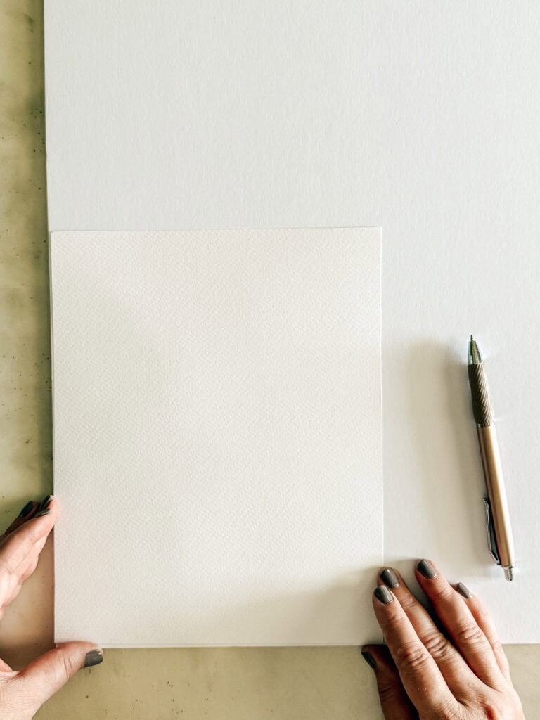 A person with gray-painted nails holds a blank sheet of textured white paper on a larger white surface. A pen lies on the right side of the paper.