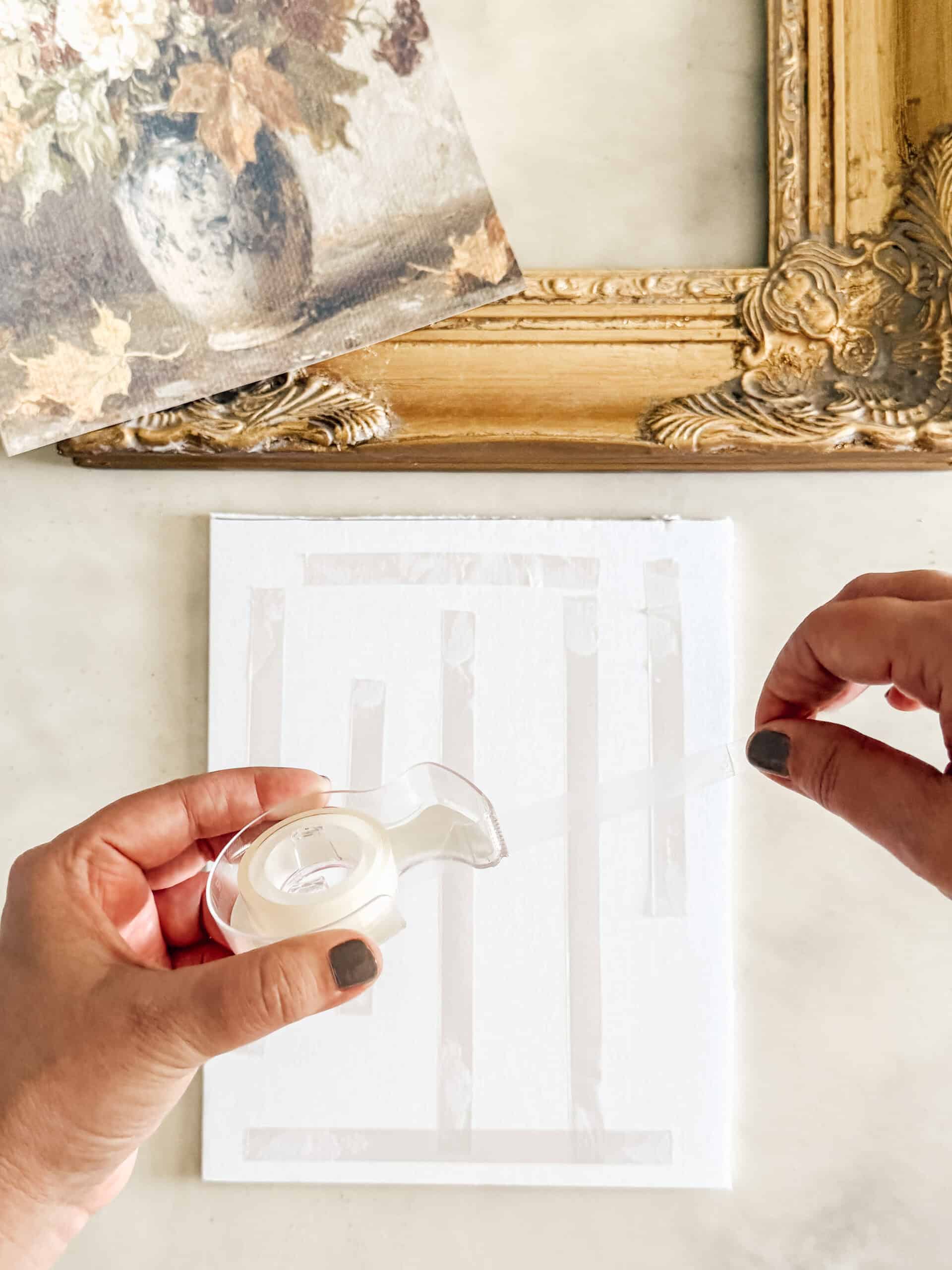 A person applies strips of clear tape to a white canvas using a tape dispenser, with a decorative gold picture frame and a floral painting visible in the background.