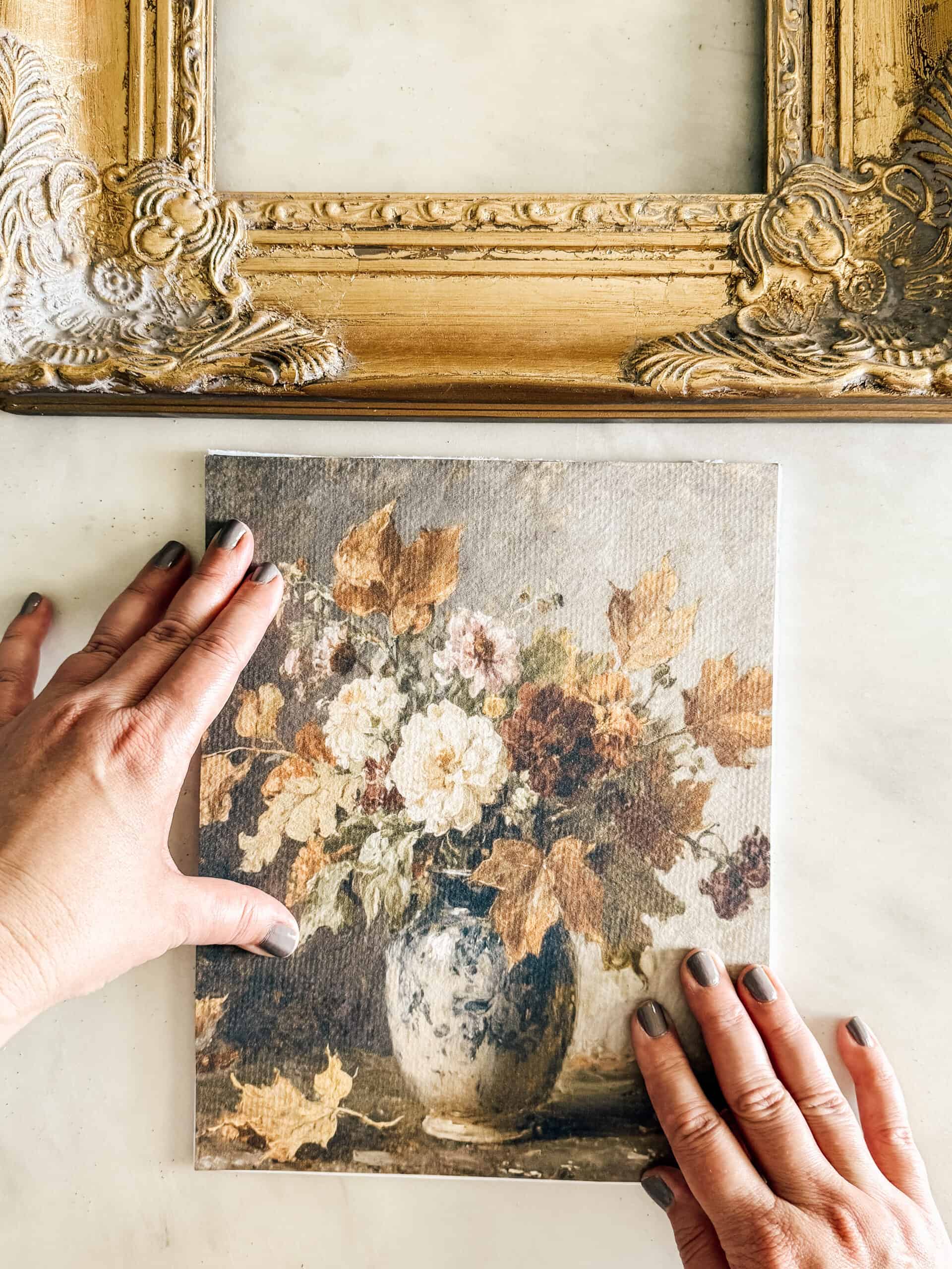 Two hands hold a small painting of a floral arrangement in a vase, with autumn leaves and roses, on a white surface. An ornate, empty gold picture frame is placed above the painting.