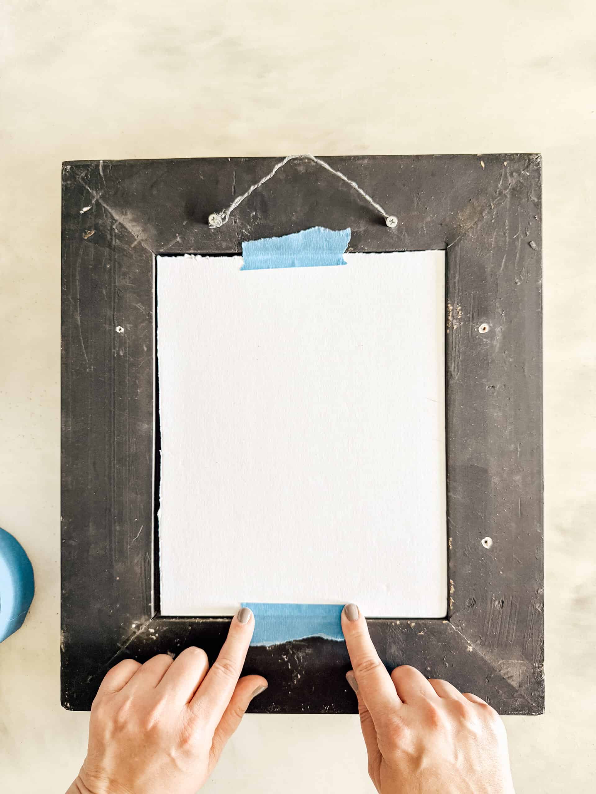 A person’s hands taping a white sheet of paper onto the back of a black picture frame using blue painter’s tape. The frame is lying flat on a light surface.