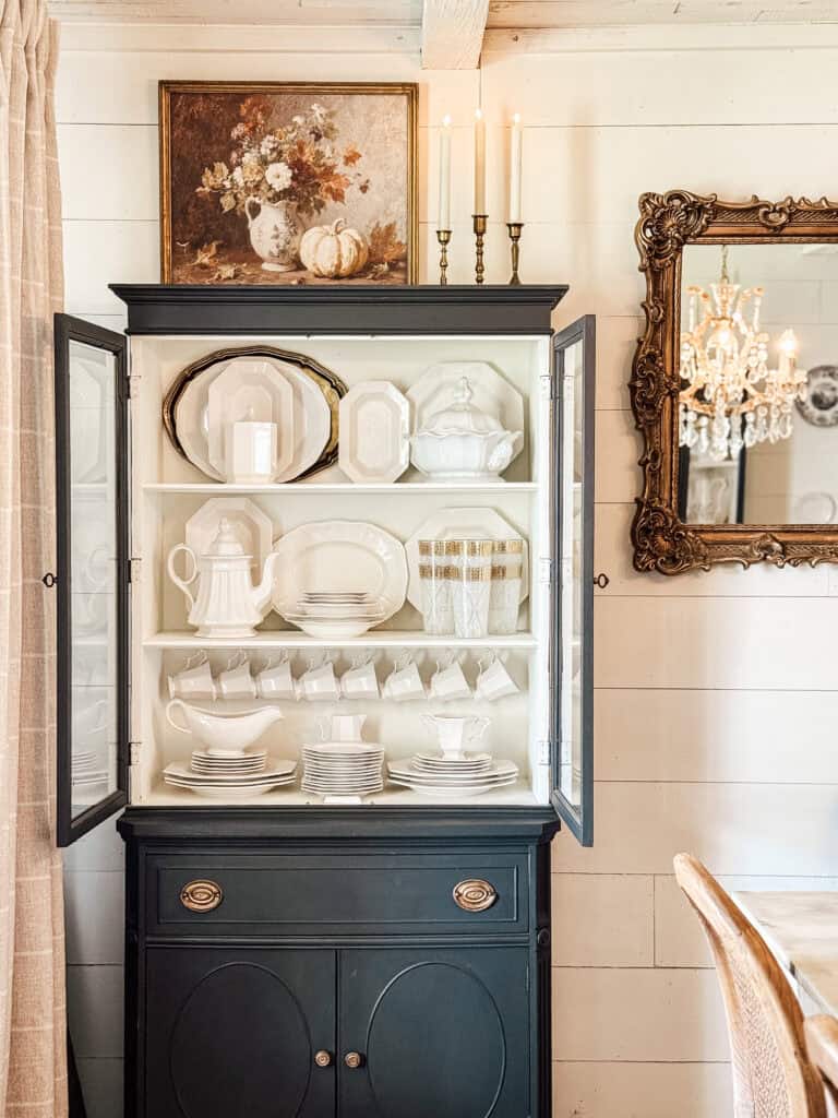 A black and white china cabinet with glass doors displays neatly arranged white dishes and teacups. Above it hangs a floral painting and two lit candles. A gold-framed mirror and chandelier are visible on the wall nearby.