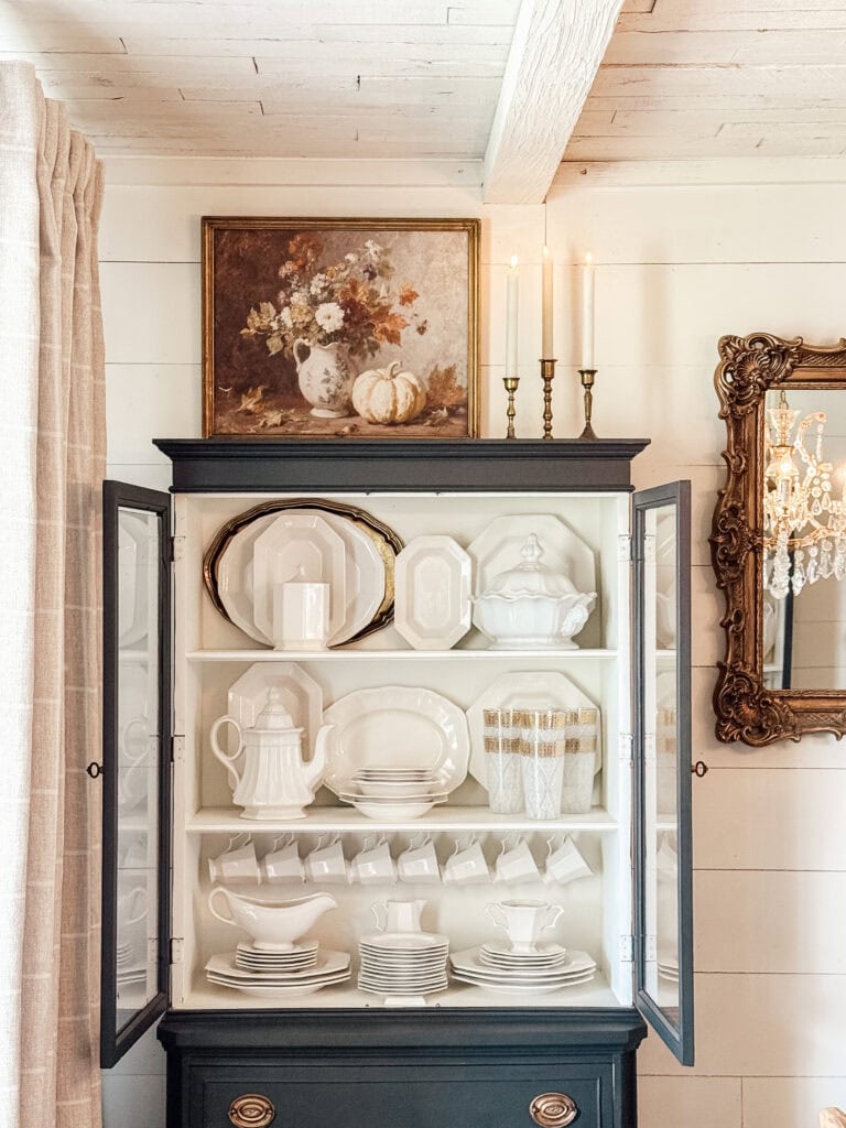A black cabinet with glass doors displays neatly arranged white china dishes, teacups, and serving pieces. On top sits a still-life painting, three tall candles, and an ornate mirror hangs on the adjacent wall.