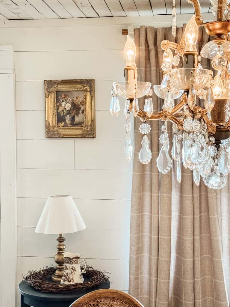 A cozy room corner with a crystal chandelier, a framed floral painting on a white shiplap wall, beige curtains, a brass lamp with a white shade on a round table, and a wicker chair partially visible.