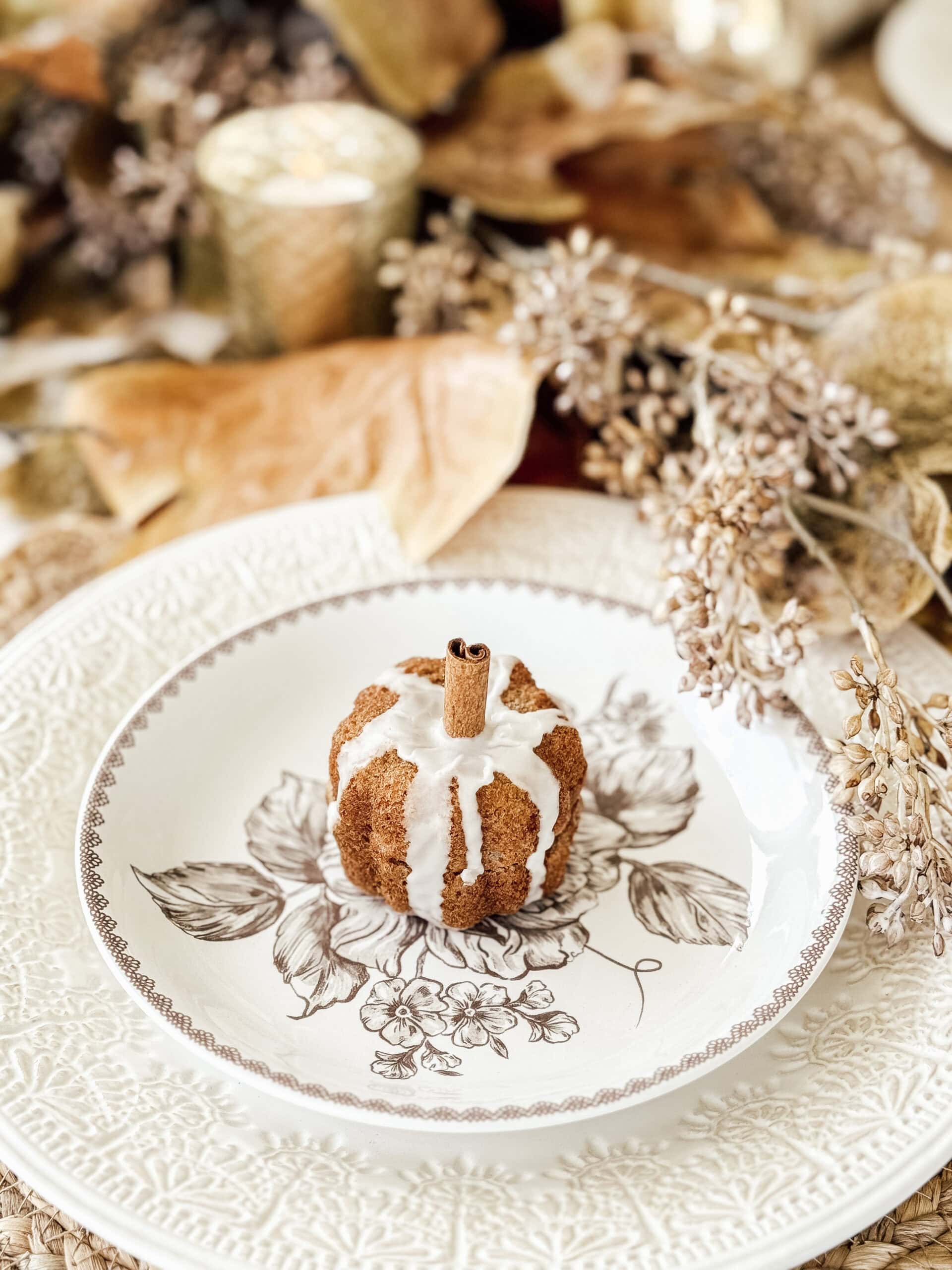 A small bundt cake with white icing and a cinnamon stick on top sits on a decorative floral plate, surrounded by autumn leaves and dried flowers, with a candle in the background.