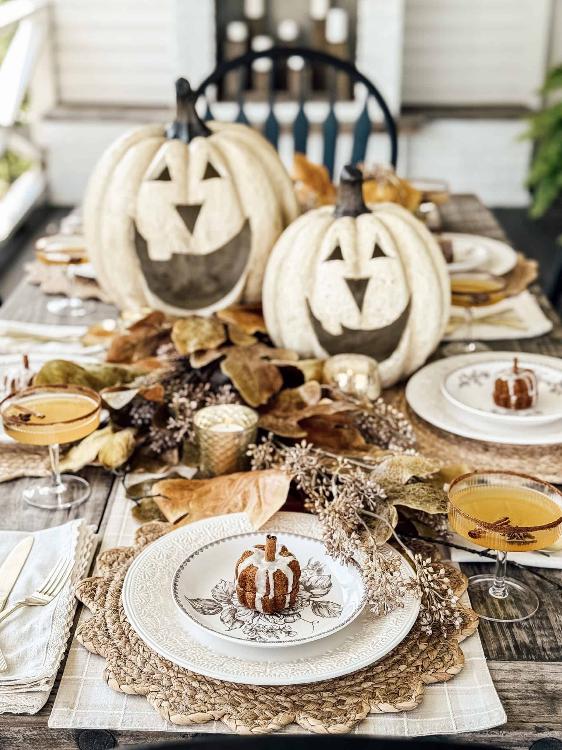 A rustic autumn table setting features white pumpkin decorations with carved faces, fall leaves, woven placemats, and small pumpkin-shaped cakes on white plates, with orange drinks in coupe glasses.