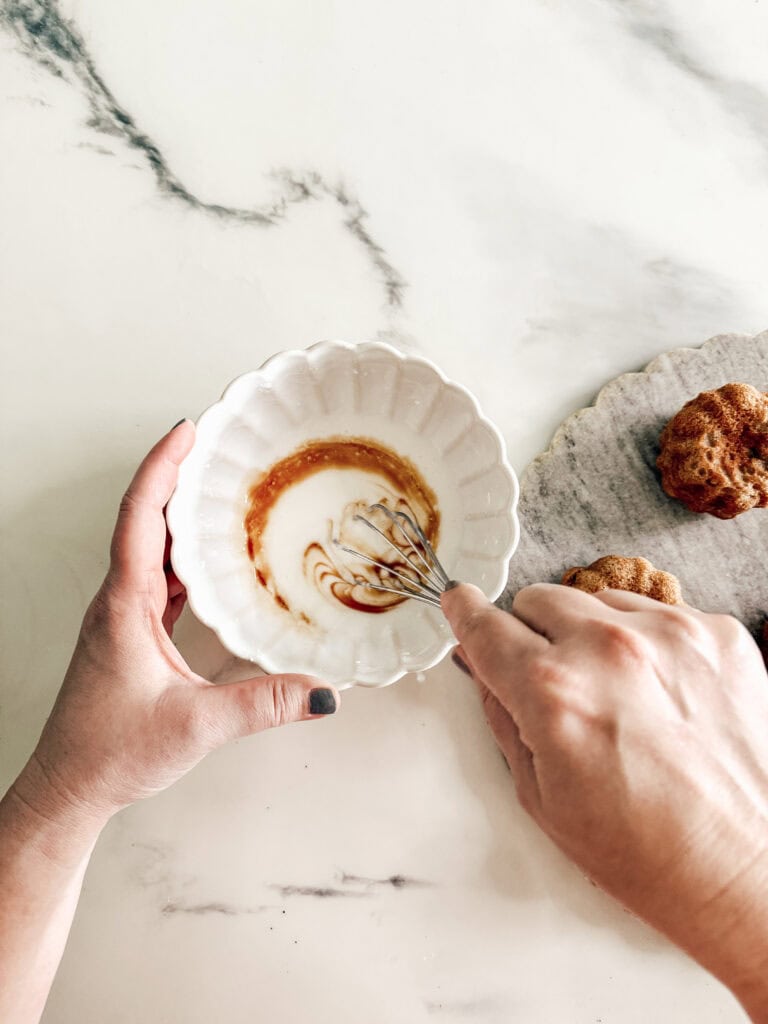 A person whisks icing in a white bowl on a marble surface, with a few baked goods on a gray tray nearby.