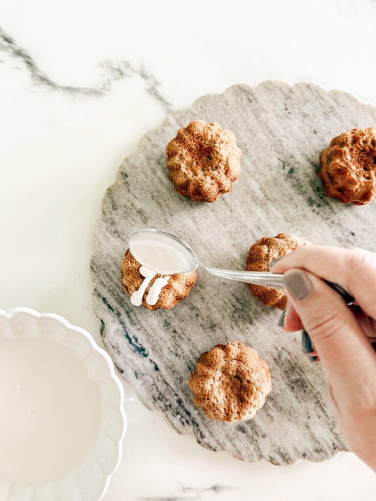 A hand holding a spoon drizzles white icing onto small, round baked cakes placed on a gray marble platter. A bowl of icing sits nearby on a white surface.
