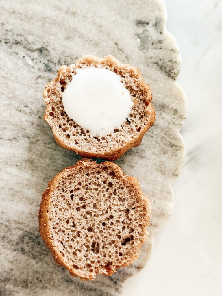 A muffin sliced in half sits on a scalloped marble surface. The top half shows a white creamy filling in the center, while the bottom half displays a light, airy crumb.