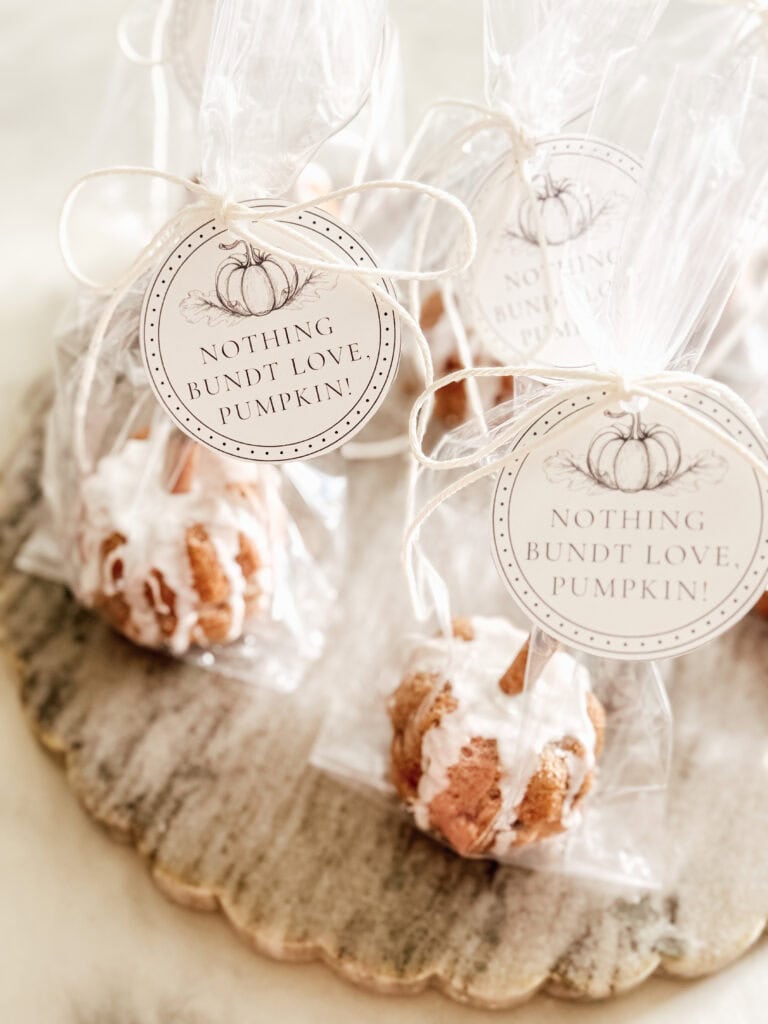 Individually wrapped mini bundt cakes with white icing sit on a wooden tray. Each package has a round tag with a pumpkin illustration and the text, Nothing bundt love, pumpkin!.