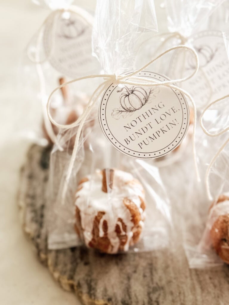 Mini pumpkin-shaped bundt cakes, drizzled with icing, are wrapped in clear bags tied with twine and labeled with tags reading “Nothing Bundt Love, Pumpkin.” The cakes are displayed on a rustic wooden surface.