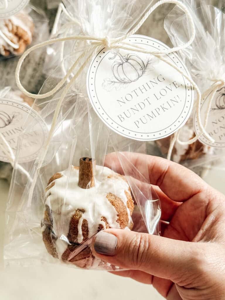 A hand holds a mini pumpkin-shaped bundt cake with white icing, wrapped in clear plastic and tied with twine. A round tag reads, “NOTHING BUNDT LOVE, PUMPKIN!” Other wrapped cakes are in the background.