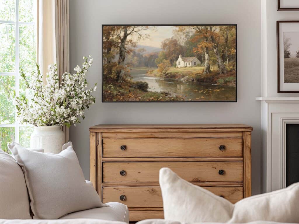 A cozy living room with a wooden dresser, a white sofa with cushions, a vase of white flowers, and a framed digital vintage art landscape painting of a cottage by a lake surrounded by autumn trees hanging on the wall.