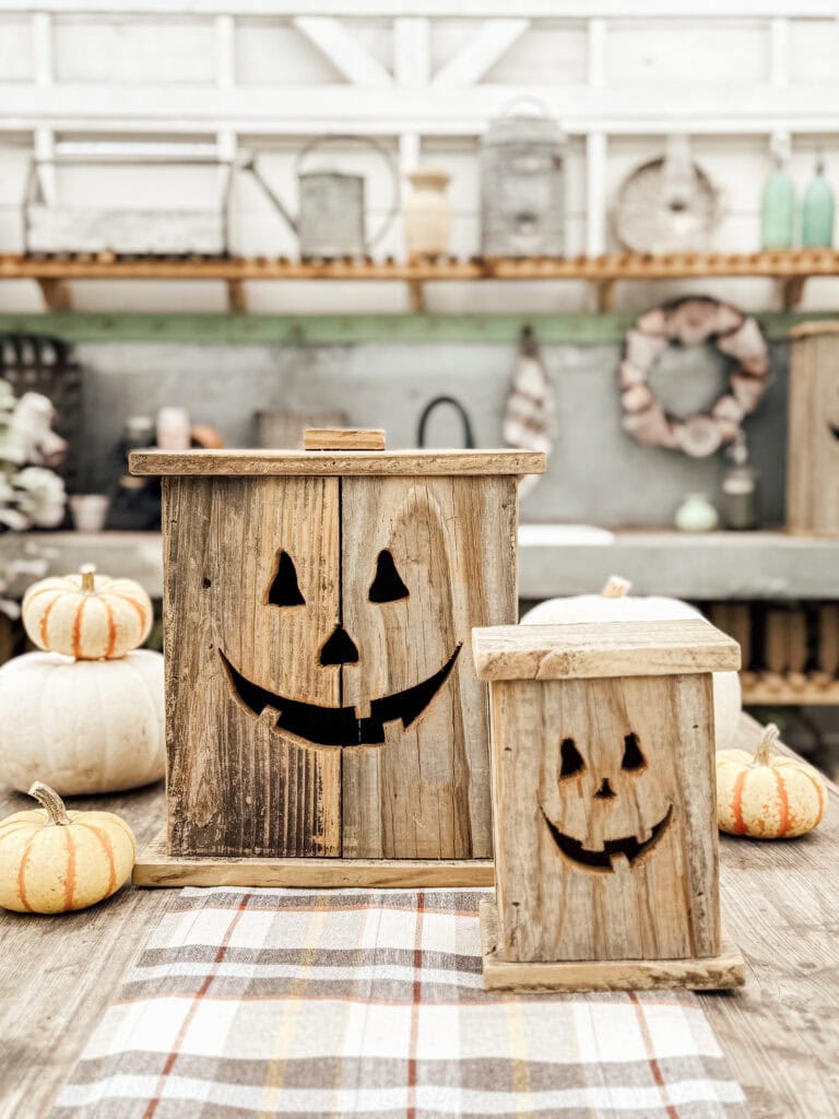 Two rustic wooden boxes with carved jack-o’-lantern faces sit on a plaid cloth, surrounded by small pumpkins, in a cozy farmhouse kitchen decorated for autumn.
