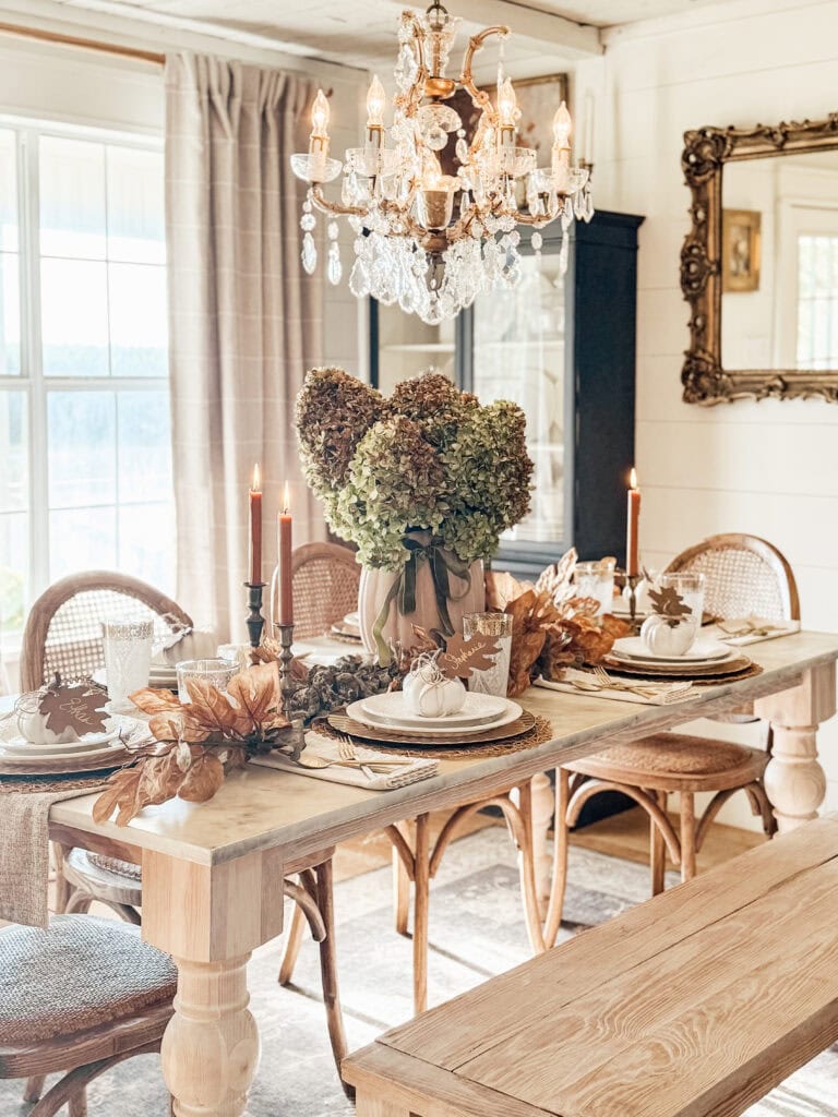A rustic dining table is set for fall with a centerpiece of hydrangeas, candles, white pumpkins, and autumn leaves, surrounded by wooden chairs and a bench under an ornate chandelier in a bright, elegant room.