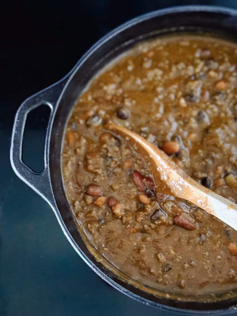 A close-up of a pot filled with thick, brown bean chili being stirred with a wooden spoon. The chili contains visible beans and is simmering in a dark, round cast iron pot.