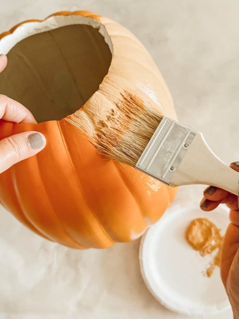 A person uses a paintbrush to apply light brown paint to an artificial orange pumpkin, with a white paint palette visible nearby.