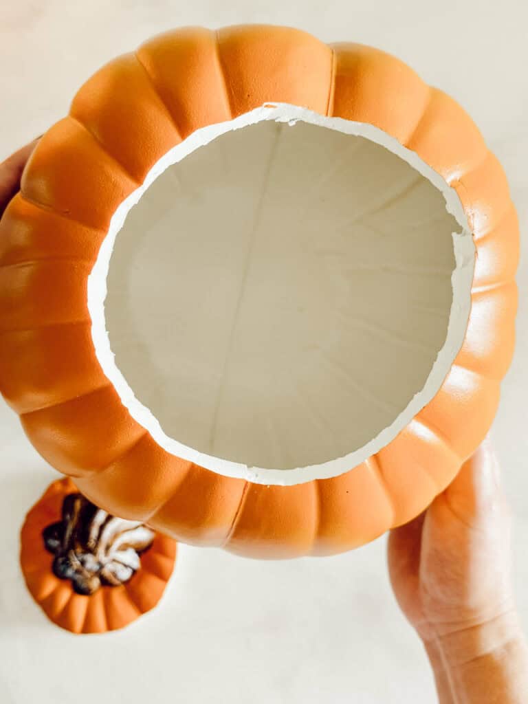A hand holds a hollow, orange pumpkin with the top cut off, revealing its empty, white interior. The cut lid with a brown stem rests nearby on a light surface.