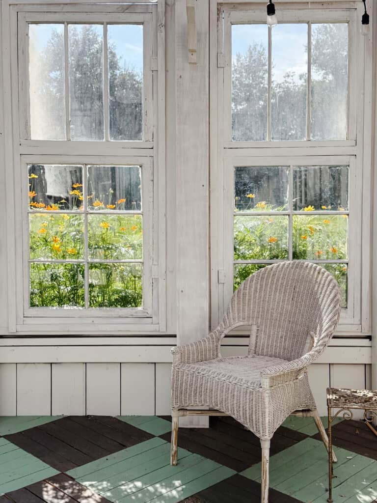 A white wicker chair sits in a sunlit room with large windows, overlooking a garden with green grass and blooming yellow-orange flowers on a bright day.